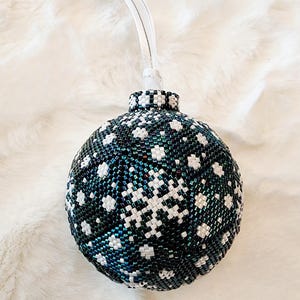 May include: A beaded Christmas ornament with a snowflake design. The ornament is primarily dark teal with white snowflake patterns. It has a silver-colored top and a white ribbon for hanging. The ornament is approximately 3 inches in diameter.