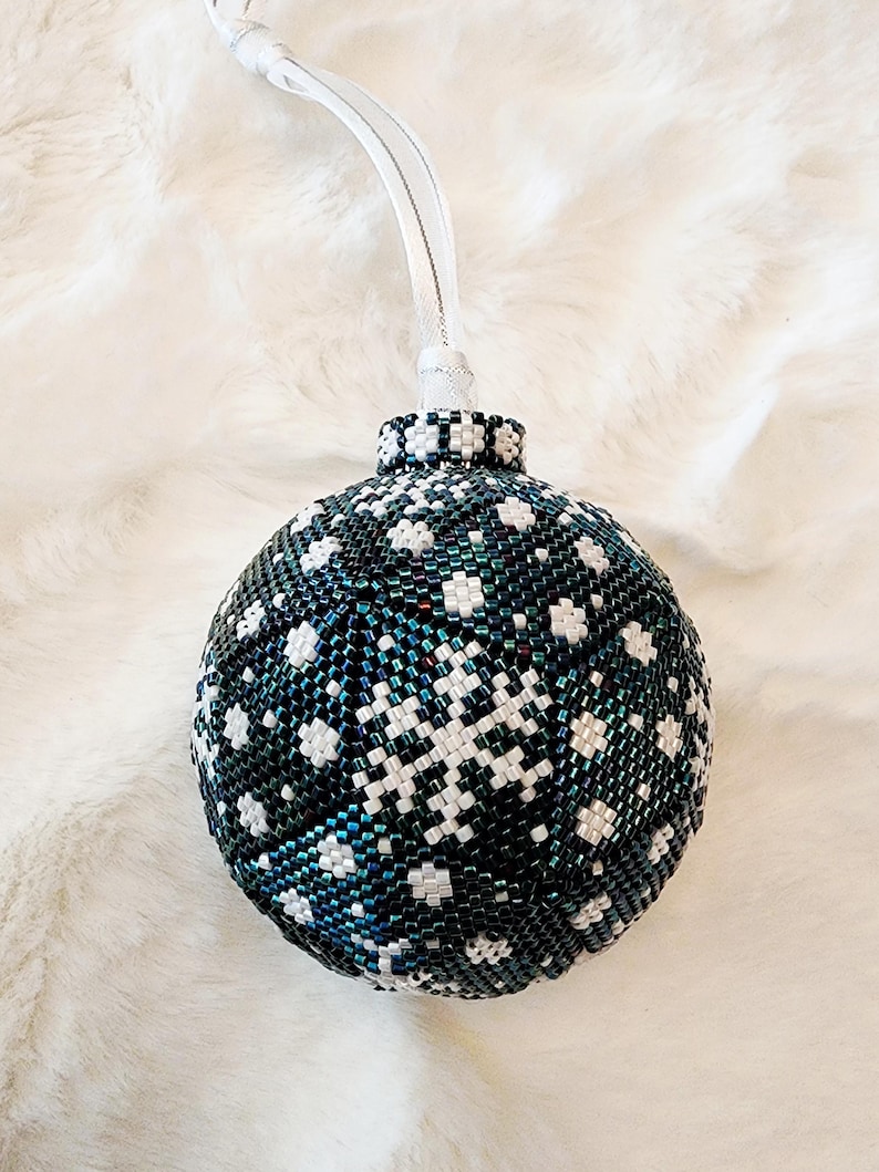 May include: A beaded Christmas ornament with a snowflake design. The ornament is primarily dark teal with white snowflake patterns. It has a silver-colored top and a white ribbon for hanging. The ornament is approximately 3 inches in diameter.