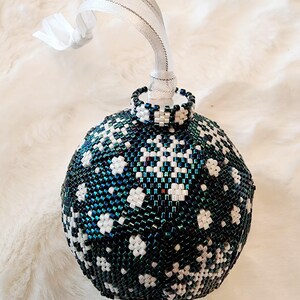 May include: A beaded Christmas ornament with a snowflake design. The ornament is primarily dark teal and white, with a white ribbon for hanging. The ornament is approximately 3 inches in diameter.