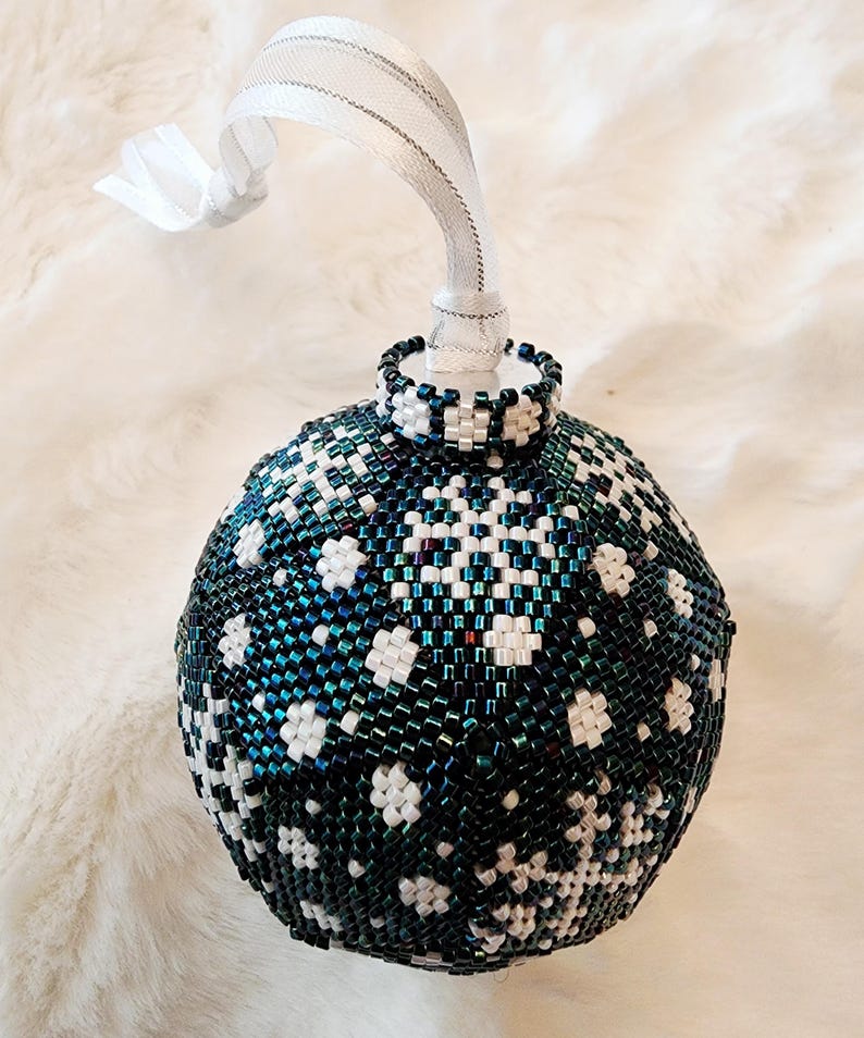 May include: A beaded Christmas ornament with a snowflake design. The ornament is primarily dark teal and white, with a white ribbon for hanging. The ornament is approximately 3 inches in diameter.