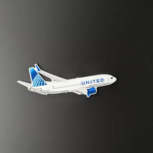 May include: A white and blue model aeroplane with the word "UNITED" printed on the side. The model features the United Airlines logo on the tail and wings. The aeroplane is set against a black background.