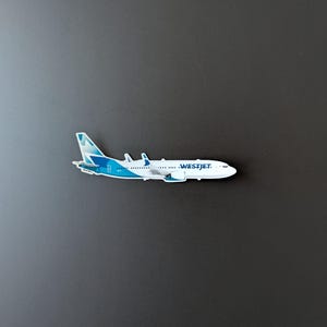 May include: A white and blue airplane magnet with the text "WESTJET" on the side. The airplane has blue and white accents on the tail and wings. The magnet is on a dark grey surface.