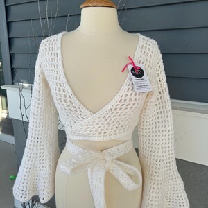 May include: A cream-colored, open-weave crochet wrap top with long, flared sleeves and a tie closure. The top is displayed on a mannequin. A tag with the text "MAY" is attached to the top.