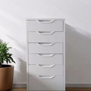 May include: A white six-drawer chest of drawers with curved white handles. The unit stands against a white wall, with a potted plant in a brown pot to the left. The drawers are evenly spaced.