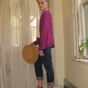 May include: A woman wearing a magenta top, blue jeans, and brown platform sandals. She is holding a straw hat in her left hand.