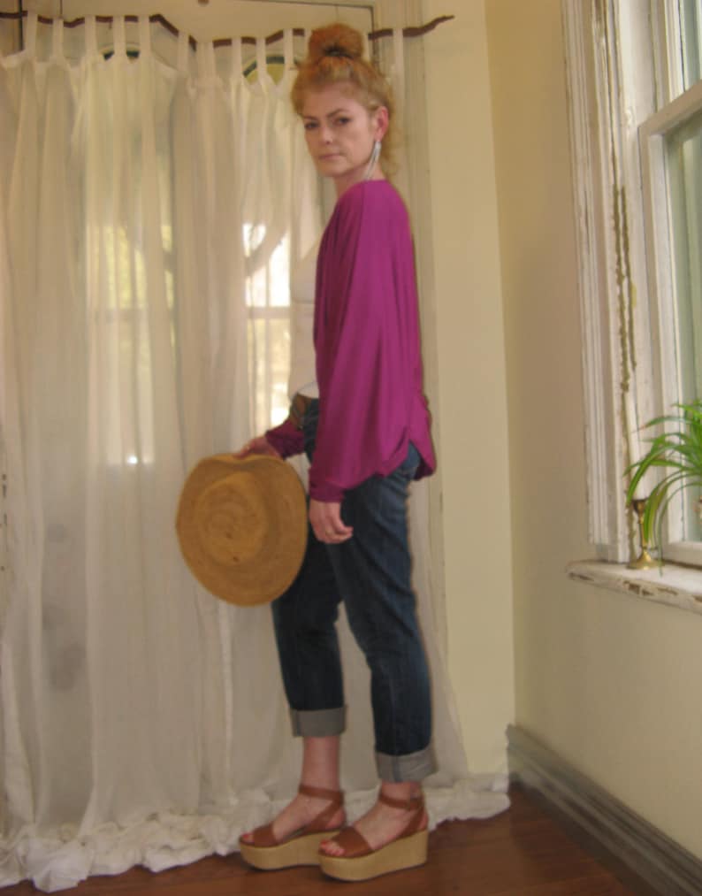 May include: A woman wearing a magenta top, blue jeans, and brown platform sandals. She is holding a straw hat in her left hand.