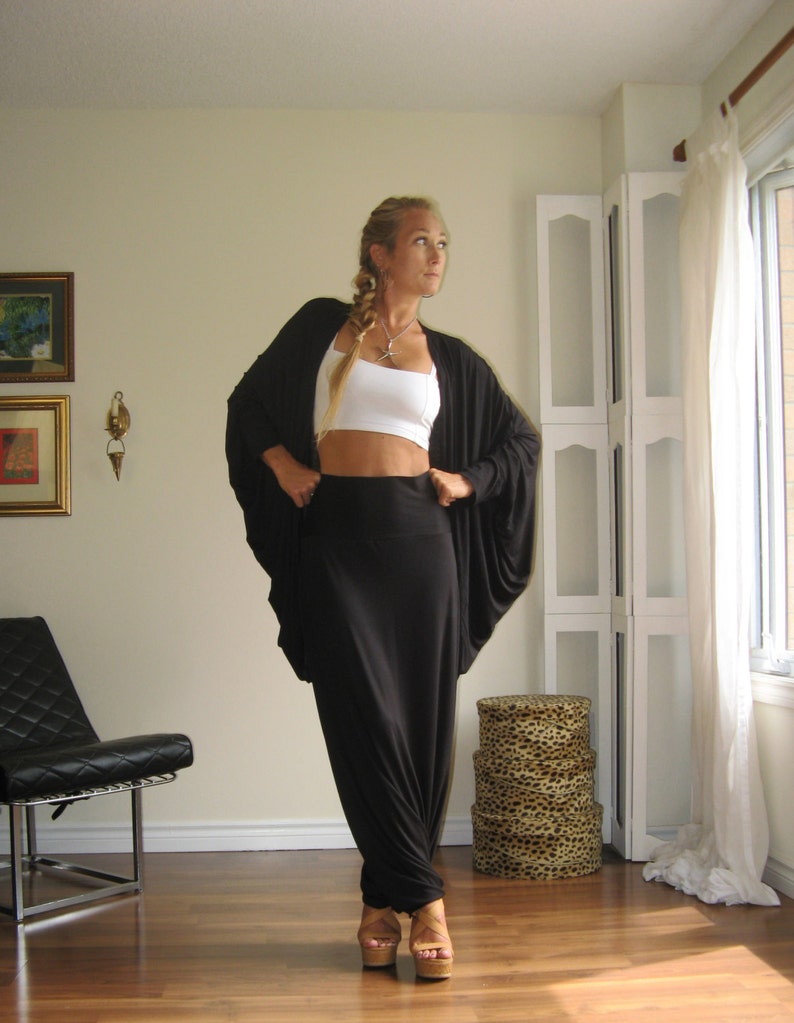 May include: A woman wearing a black long-sleeved top with a draped design and a black maxi skirt. She is wearing a white tank top underneath and has a silver necklace. She is standing in a room with a white wall and a window in the background.