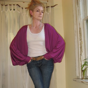 May include: A woman wearing a purple cardigan with long sleeves and a white tank top. She is wearing blue jeans and a brown belt.
