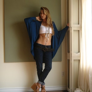 May include: A woman wearing a blue long-sleeved shirt with a white tank top underneath. She is wearing a silver necklace with feathers, a brown belt, and blue jeans. She is standing in front of a white door with a green wall behind her.