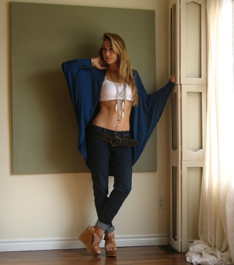 May include: A woman wearing a blue long-sleeved shirt with a white tank top underneath. She is wearing a silver necklace with feathers, a brown belt, and blue jeans. She is standing in front of a white door with a green wall behind her.