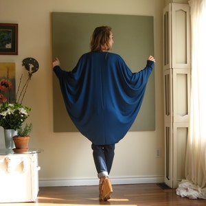 May include: A woman wearing a long, flowing, royal blue tunic top with a wide, loose fit. The top has a scoop neckline and long sleeves. The woman is wearing blue jeans and brown heeled sandals.