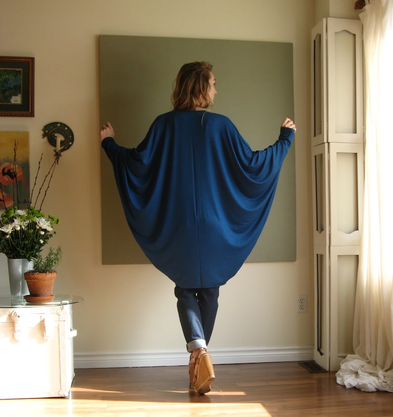 May include: A woman wearing a long, flowing, royal blue tunic top with a wide, loose fit. The top has a scoop neckline and long sleeves. The woman is wearing blue jeans and brown heeled sandals.