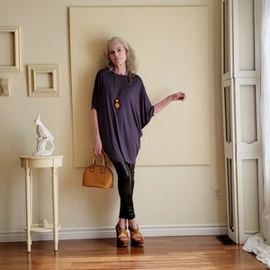 May include: A woman wearing a purple, loose-fitting top with a brown pendant necklace, black leather trousers, and brown platform sandals. She is holding a brown leather handbag.