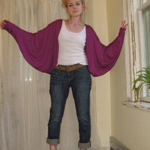 May include: A woman wearing a purple, long-sleeved cardigan with a white tank top underneath. She is wearing blue jeans and brown sandals.