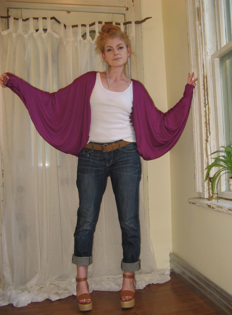 May include: A woman wearing a purple, long-sleeved cardigan with a white tank top underneath. She is wearing blue jeans and brown sandals.