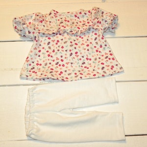 May include: A white baby outfit with a floral print top and white trousers. The top has a ruffled neckline and short sleeves.