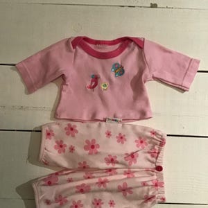 以下が含まれることがあります： ピンク色のベビー服セット。長袖トップスとパンツが含まれています。トップスはピンク色のトリムと鳥と蝶の刺繍デザインが施されています。パンツにはピンクの花柄がプリントされています。柔らかい素材で作られています。