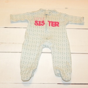 Puede incluir: Un pijama de bebé azul claro con un estampado floral y las palabras "LITTLE SIS TER" en rosa. El pijama tiene una cremallera en la parte delantera y pies.
