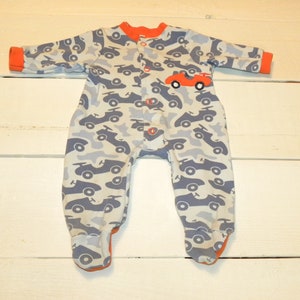 May include: A blue and white baby sleepsuit with a red racing car design. The sleepsuit has a popper fastening at the crotch and a button fastening at the neck.