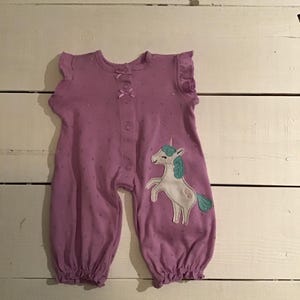 Könnte beinhalten: Lilafarbener Baby-Strampler mit einem Einhorn-Applikation. Der Strampler hat einen Druckknopfverschluss und Rüschenärmel.