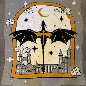 May include: A rectangular crochet blanket featuring a central arched window design with a black dragon silhouette, a yellow crescent moon, and white stars. The background is light gray, and the bottom shows a gray castle with white clouds.