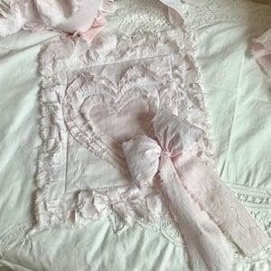 May include: A set of pale pink bedding with ruffled edges and a heart-shaped appliqué. The set includes a pillow, a ruffled bed skirt, and a square decorative piece with a bow.