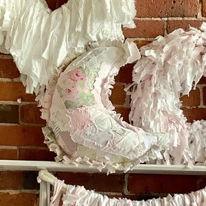 May include: A crescent moon-shaped fabric pillow featuring a patchwork design of pink roses, white floral patterns, and light pink fabric scraps. The edges are finished with frayed fabric strips, creating a textured, layered appearance.