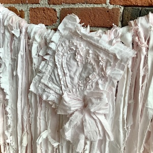 May include: A pale pink fabric garland featuring long, frayed strips of cloth hanging vertically. A central square fabric panel displays a ruffled heart shape in the middle, accented with a large, tied fabric bow at the bottom edge.