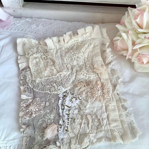 May include: A square, off-white decorative pillow with a patchwork design. The pillow features various lace and fabric textures, including floral appliqués and ruffled edges. A small piece of fabric has text printed on it. The pillow is on a white surface.