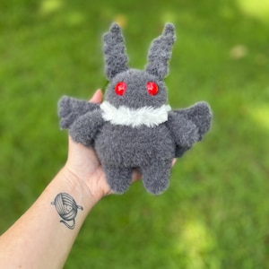 May include: A handmade gray plush moth toy with red eyes and a white collar. The moth has small wings and two upright antennae. The toy is made of a soft, fuzzy material. The background is blurred green grass.