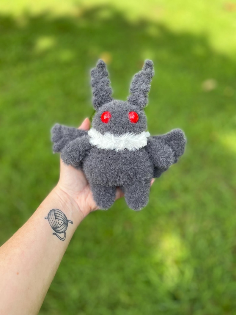 May include: A handmade gray plush moth toy with red eyes and a white collar. The moth has small wings and two upright antennae. The toy is made of a soft, fuzzy material. The background is blurred green grass.