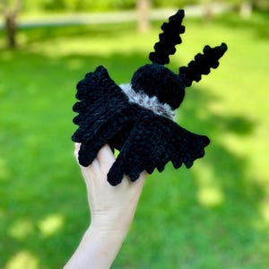 May include: A handmade black crochet moth with detailed wings and antennae. The moth has a fluffy, light-colored collar. The plush toy is held against a blurred green background, suggesting an outdoor setting. The moth is a unique, handcrafted item.