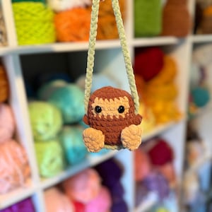 May include: A handmade crochet owl ornament in shades of brown and tan, with black eyes and a light green strap. The owl is suspended in front of a shelf filled with colorful yarn skeins, suggesting a craft or hobby theme.