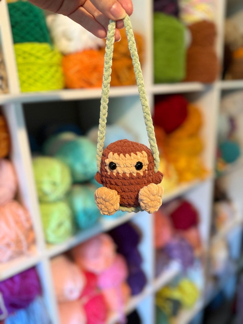 May include: A handmade crochet owl ornament in shades of brown and tan, with black eyes and a light green strap. The owl is suspended in front of a shelf filled with colorful yarn skeins, suggesting a craft or hobby theme.
