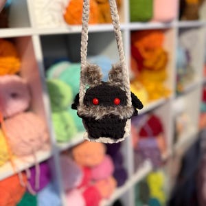 May include: A handmade black crochet bag with a furry face and red eyes. The bag has a gray strap and is held up against a backdrop of colorful yarn skeins. The bag's face has two furry ears and a white trim.