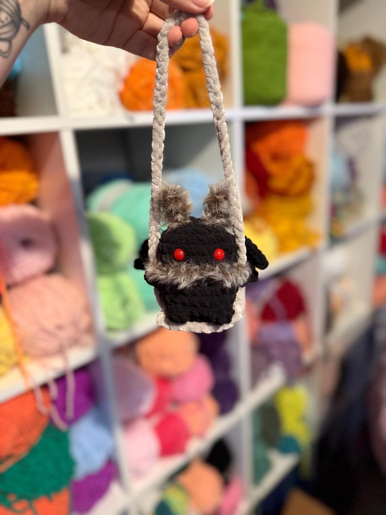May include: A handmade black crochet bag with a furry face and red eyes. The bag has a gray strap and is held up against a backdrop of colorful yarn skeins. The bag's face has two furry ears and a white trim.