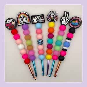 May include: A collection of colorful crochet hooks with decorative toppers. Each hook features a series of vibrant beads in various colors, leading to a metal hook. The toppers display phrases like "Mama Mommy Ma" and "Emotional Dumpster Fire."