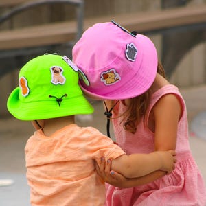 May include: Two children wearing colorful bucket hats with patches. The child on the left is wearing a green hat with a black strap and the child on the right is wearing a pink hat with a black strap. Both children are smiling and looking at the camera.