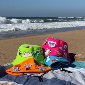 May include: Three colorful bucket hats with various colorful patches and designs on a striped beach towel. The hats are green, pink, and orange.