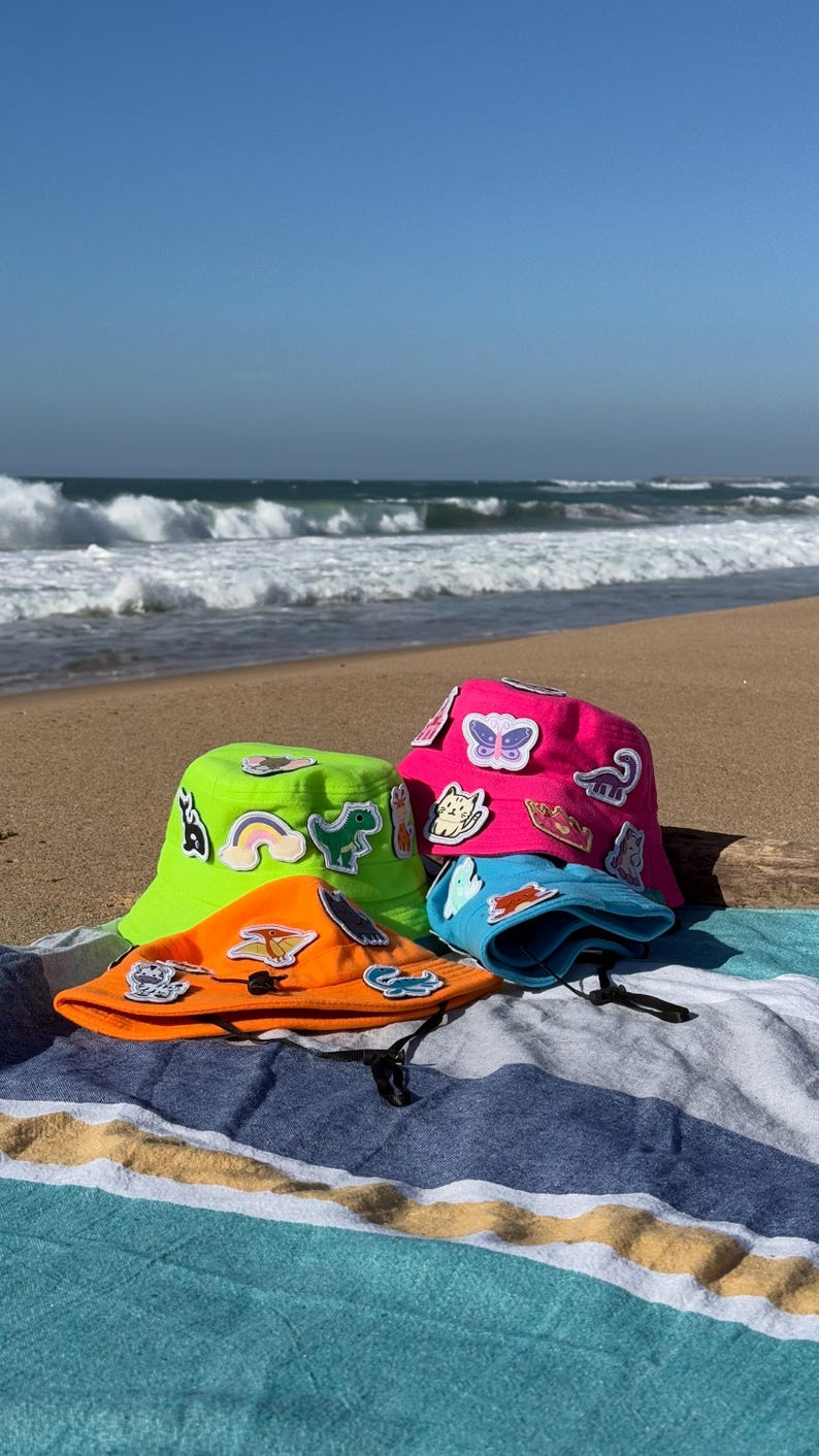 May include: Three colorful bucket hats with various colorful patches and designs on a striped beach towel. The hats are green, pink, and orange.