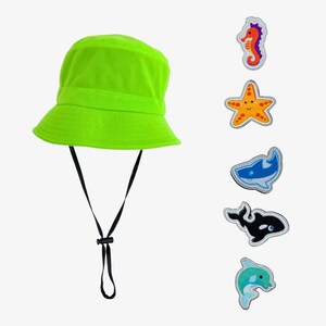 May include: A bright green bucket hat with a black adjustable strap. The hat is decorated with five colorful iron-on patches featuring a seahorse, starfish, shark, orca, and dolphin.