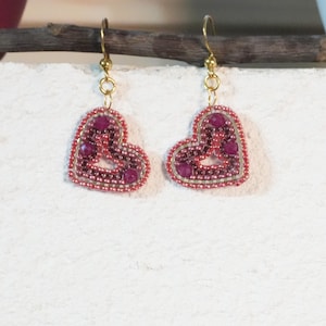May include: A pair of heart-shaped earrings. Each earring features a beaded design in shades of red and pink, with a gold-colored hook. The heart shape is outlined with small beads, and the center is filled with larger, darker red beads.