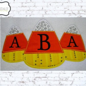 May include: Three felt candy corn shapes with orange, yellow and white stripes. Each candy corn has a letter "A" embroidered on the orange section. The candy corn shapes are on a white background.