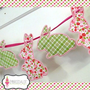 May include: A pink and green bunny garland with a pink ribbon. The bunnies are made of fabric with different patterns, including plaid and floral.