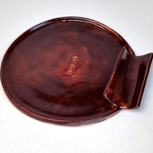 Può includere: Un posacenere rotondo in ceramica fatto a mano, con una smaltatura marrone scuro. Il posacenere presenta un appoggio curvo e la parola "GORTET" in rilievo sulla superficie. Design semplice e funzionale, adatto per la casa o l'ufficio.