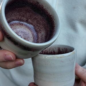 Peut inclure: Deux tasses en céramique faites à la main. Les tasses sont blanches avec un intérieur marron tacheté. Une tasse est tenue au-dessus de l'autre, mettant en valeur le design intérieur. Les tasses ont un design simple et minimaliste.