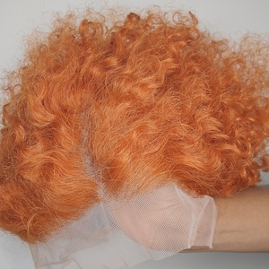 May include: A vibrant orange curly wig with a lace front. The wig features a voluminous, textured style with tightly wound curls. The lace front is a light beige color, designed to blend with the skin for a natural look. The wig is set against a plain background.