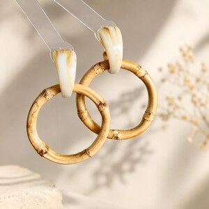 Pode incluir: Um par de brincos de argola com anéis circulares de bambu suspensos por peças de resina cor de creme lisa com um subtil marmoreado castanho. Os brincos estão presos a abas de plástico transparente que mostram a textura natural do bambu.