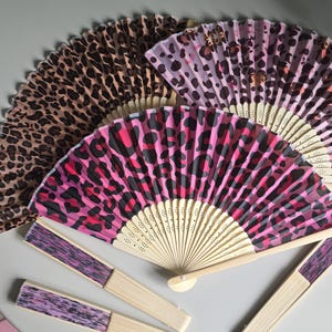 May include: Three folding hand fans with light-coloured wooden ribs and pleated fabric covers. One fan features a classic brown and black leopard print, another has a pink and black leopard pattern, and the third displays a purple and black design.
