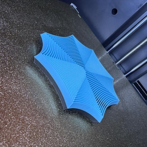 May include: A light blue, geometric-shaped object with a textured surface. The object has a series of curved lines and is set against a dark background. The design appears to be 3D printed.